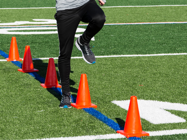 A person wearing black running shoes stepping over a row of orange cones during a practice session on a turf field with white lines.