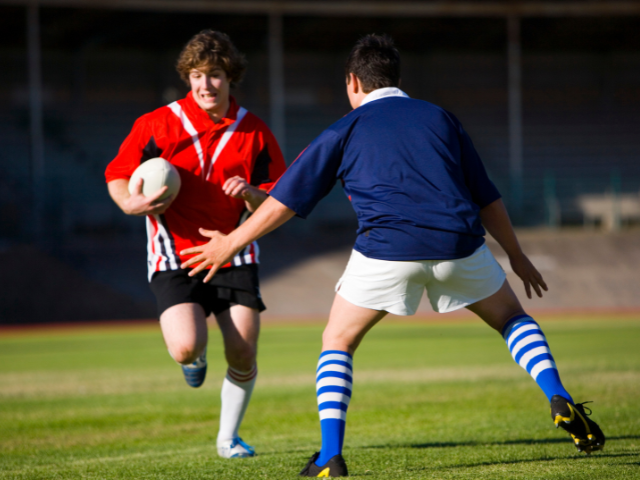 A rugby player in a red uniform holding the ball, running towards a player in a blue uniform preparing to defend.