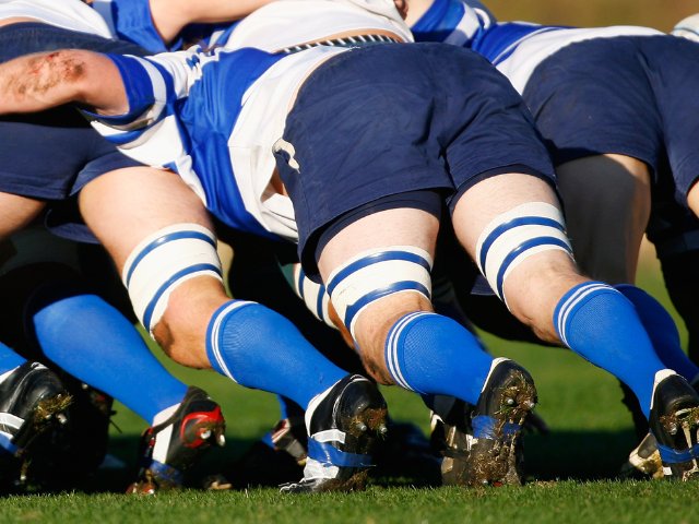 A close-up shot of rugby players in blue and white striped socks, locked in a scrum formation, digging their cleats into the ground.