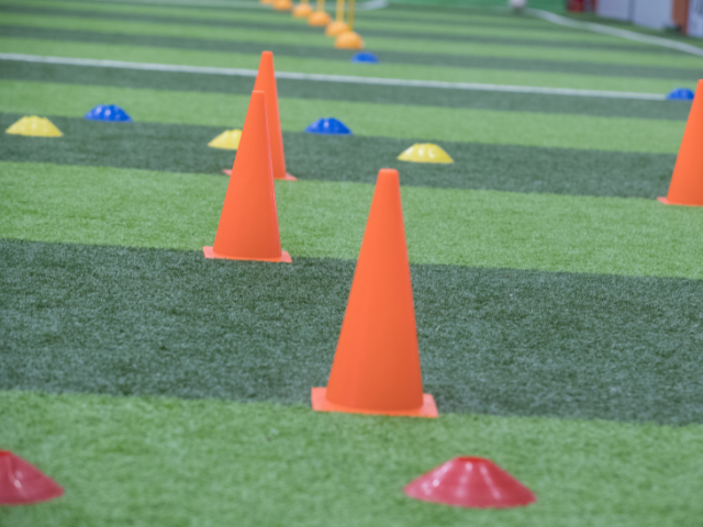 Orange cones arranged in a line on an artificial turf field with smaller colored markers in the background.