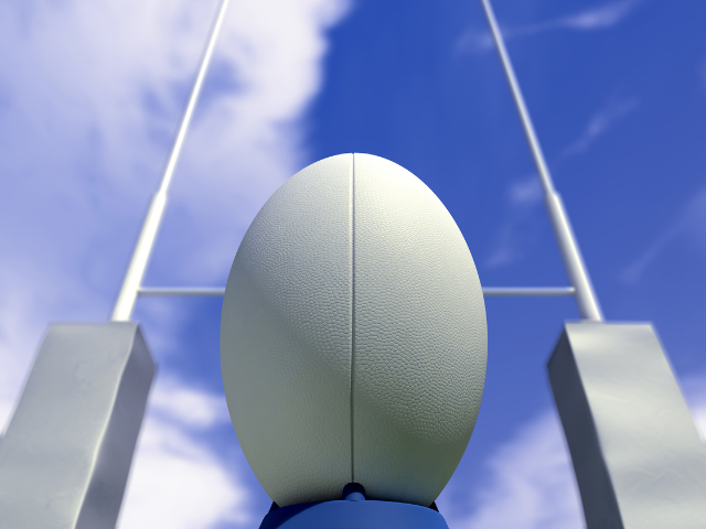 Close-up view of a rugby ball placed on a kicking tee, looking towards rugby posts under a clear sky.