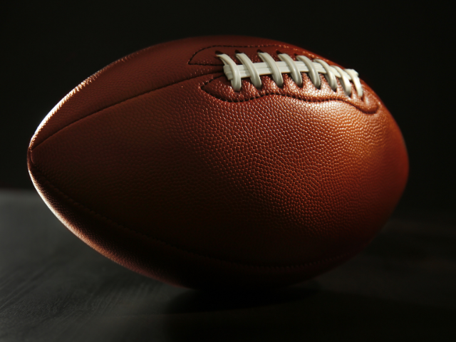 A close-up of a rugby ball on a dark background, showing the texture of the leather and laces.