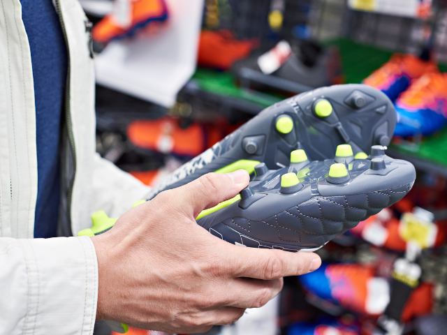 A person holding a grey rugby boot with neon yellow studs, inspecting the sole for quality.