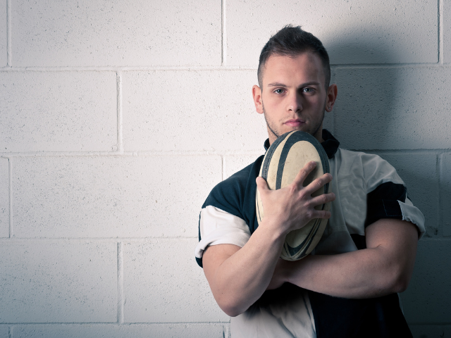 A young man in a rugby uniform holding a rugby ball against his chest.