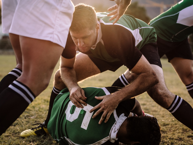 A player crouches over a teammate during a ruck, protecting the ball.