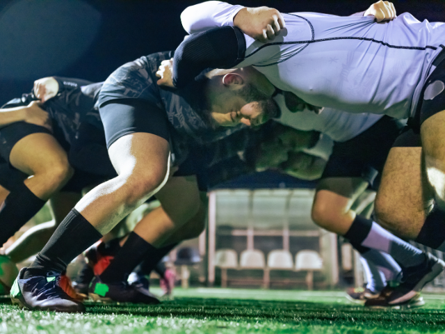 A rugby team wearing black and white jerseys crouching in a scrum formation under bright field lights during a night game.
