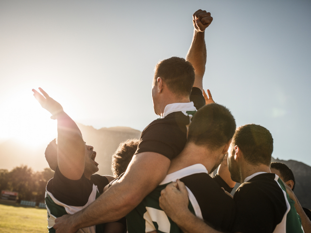 Rugby players celebrate a victory, lifting one player up while cheering as the sun sets in the background.