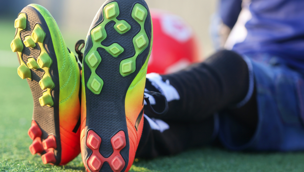 A pair of rugby boots with a mix of bright green and orange soles, showing a player sitting with feet extended.