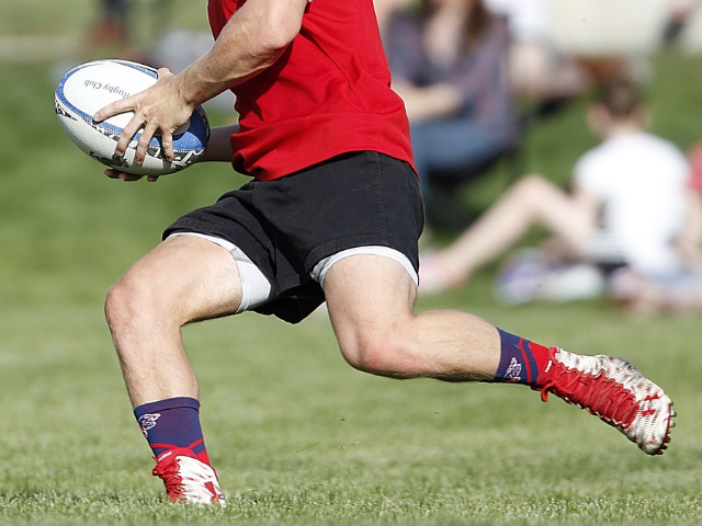 A rugby player running with a ball in red cleats during a match.